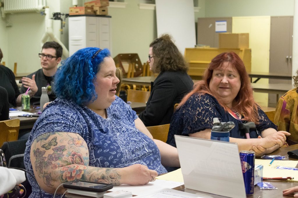 Woman at left with blue hair smiling and talking in a church hall. A woman at her right listens intently. Others in the room are talking as well.