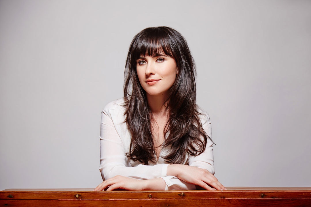 A woman with long dark brown hair and a sheer off-white long-sleeve blouse with a plunging neckline has her elbows crossed and resting on a wooden tabletop.