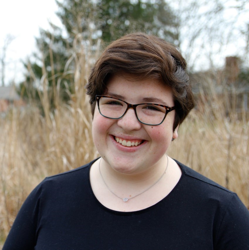 A white person with short dark brown hair, brown eyes, and freckles smiles in a straw field. They wear brown and blue glasses.