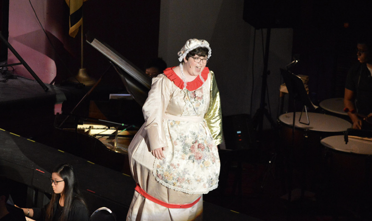 An actor with short brown hair wears a white silky bonnet and dress designed to look like a teapot.
