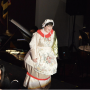 An actor with short brown hair wears a white silky bonnet and dress designed to look like a teapot.